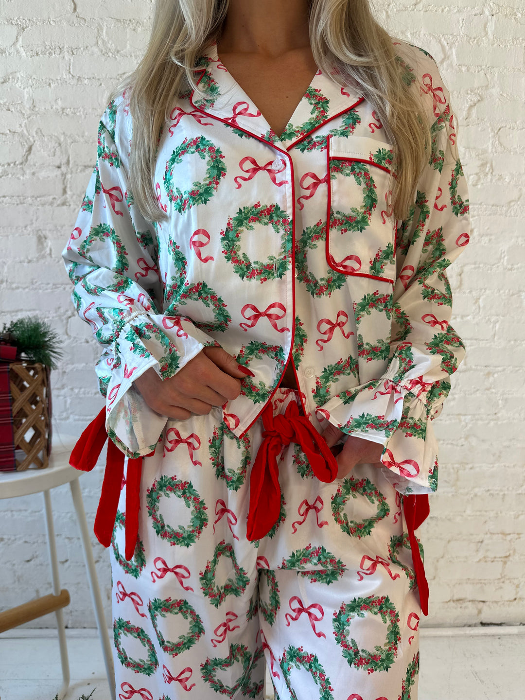 Woman wearing white satin pajama set with green wreaths all over, and red velvet trim. 