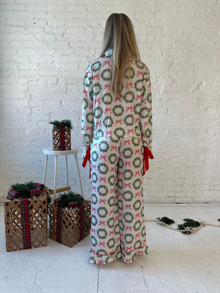 Woman wearing white satin pajama set with green wreaths all over, and red velvet trim. 