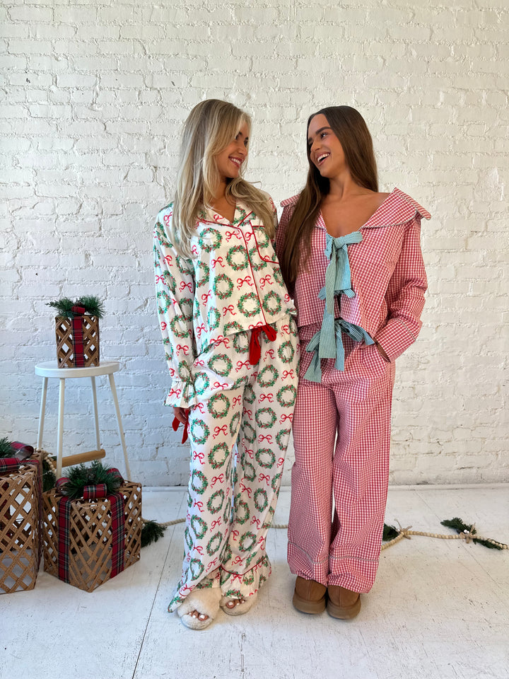 Woman wearing white satin pajama set with green wreaths all over, and red velvet trim. 