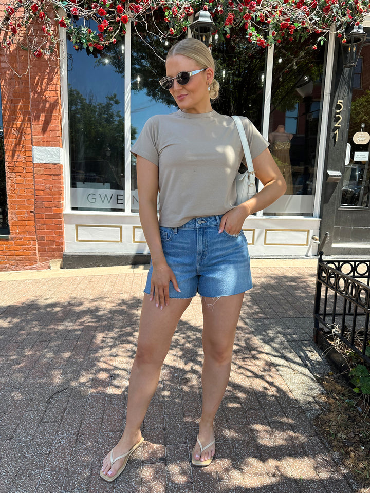 Woman wearing taupe baby tee with medium wash denim shorts. 