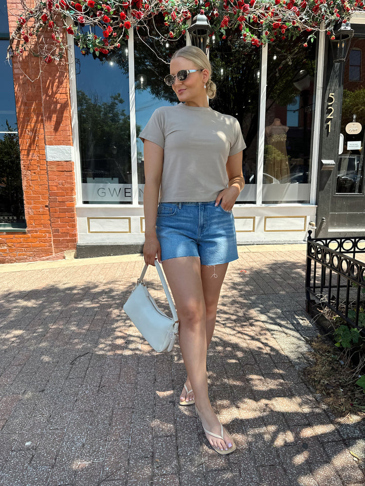 Woman wearing taupe baby tee with medium wash denim shorts. 